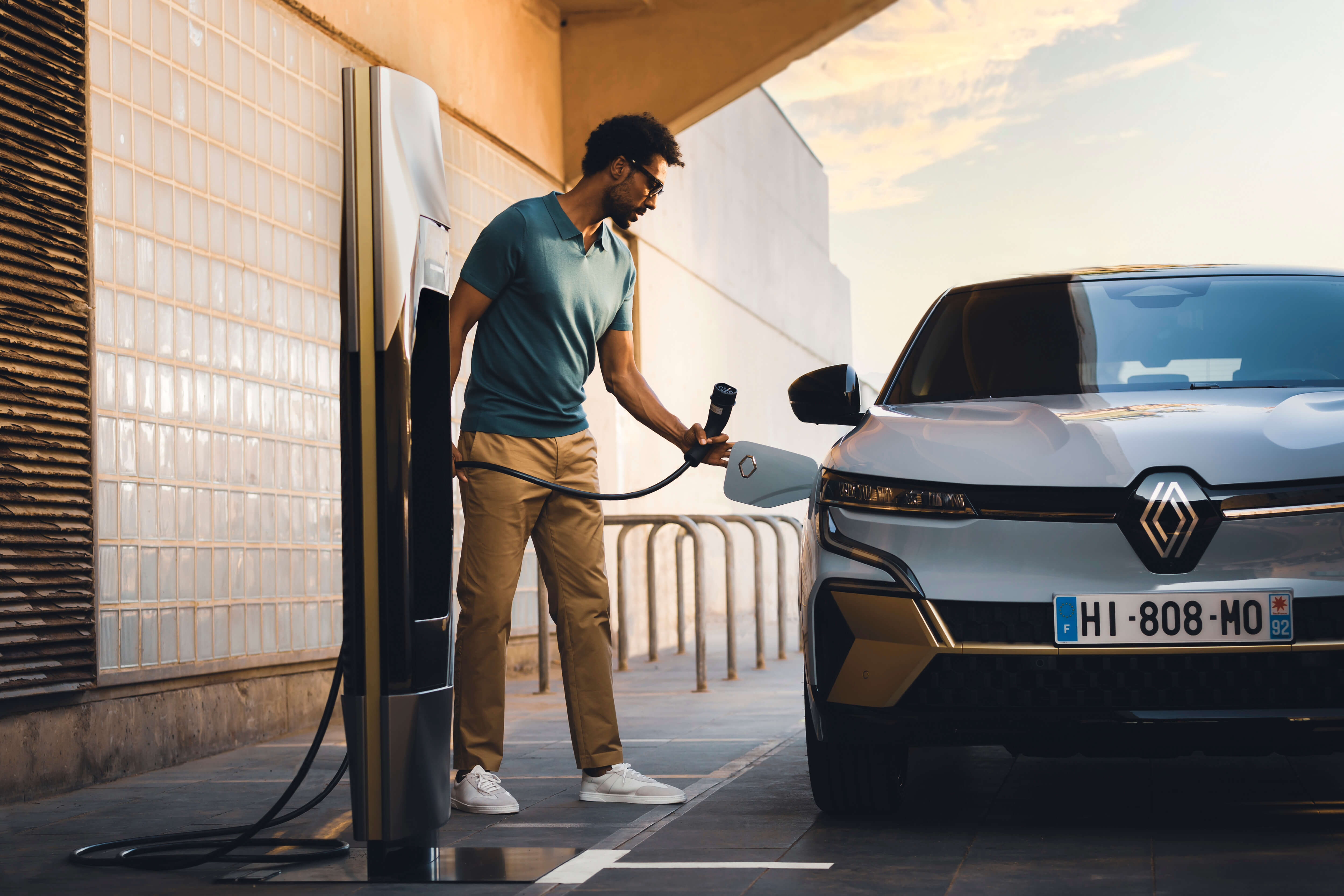 Pilkas visiškai elektrinis Renault MEGANE E-TECH Electric stovi prie įkrovimo stotelės, vyras ketina įjungti krovimo kabelį į elektromobilio krovimo jungtį