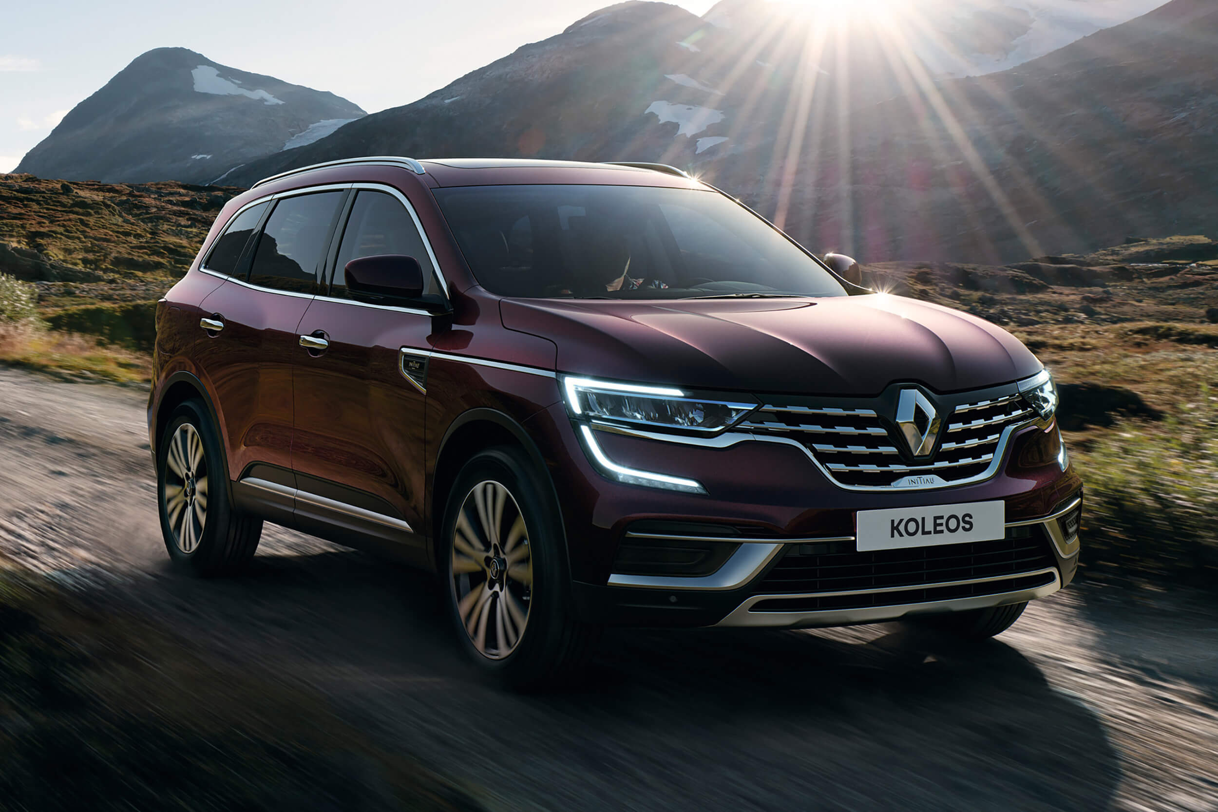 Tamsiai raudonos spalvos visureigis Renault Koleos važiuoja lauko keliu, fone matyti kalnai