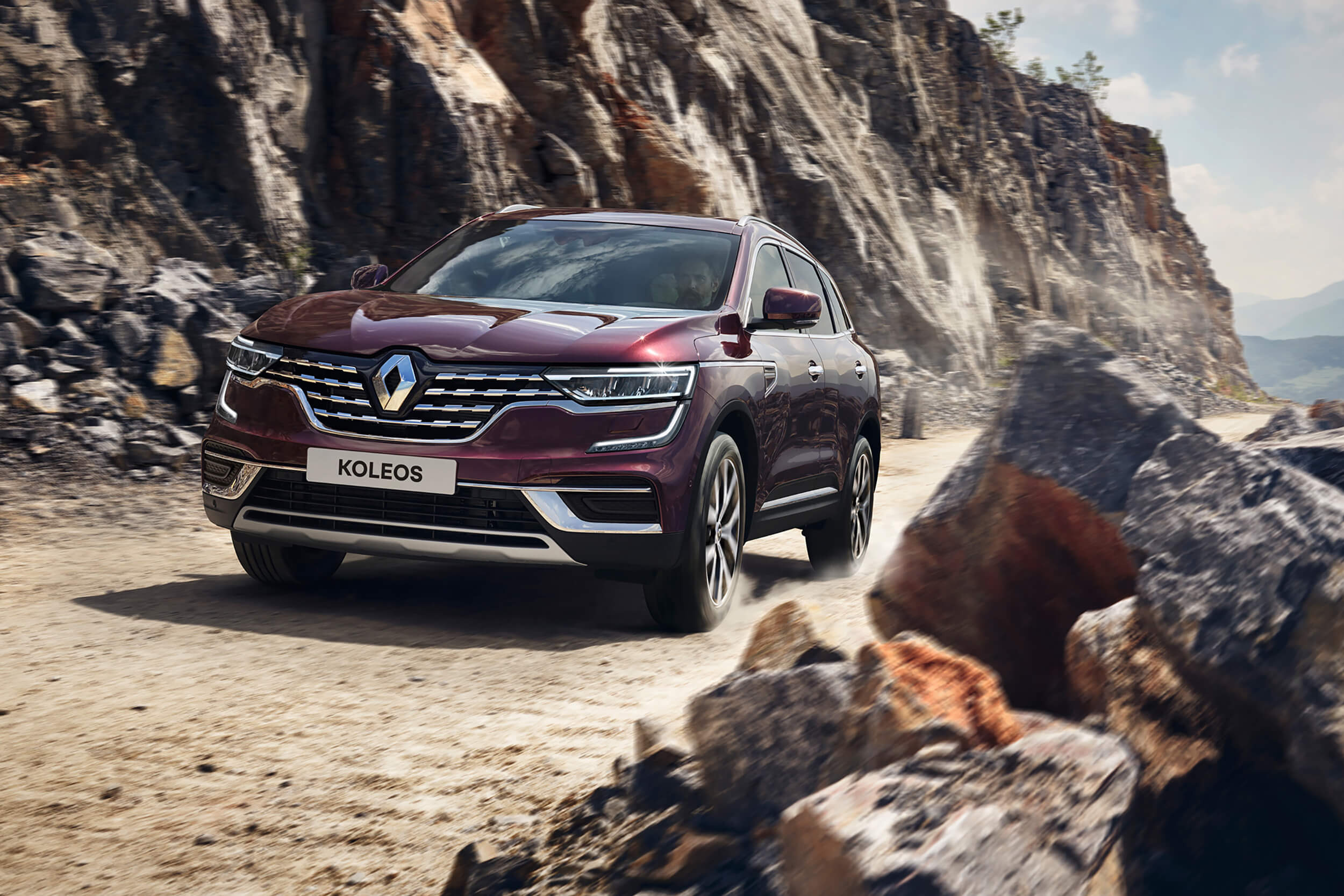 Tamsiai raudonos spalvos visureigis Renault Koleos važiuoja žvyrkeliu tarp kalnų